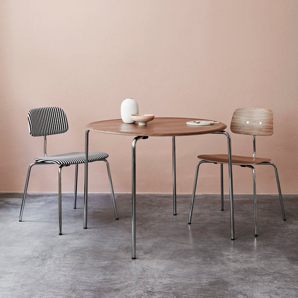 Round wooden table with light brown finish, accompanied by gray and light brown striped chairs against a soft pink backdrop.