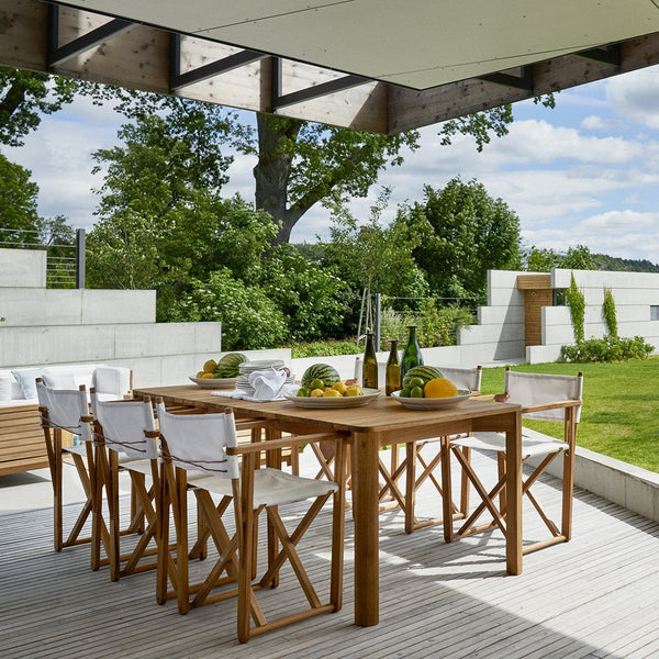 "Natural wood dining table with white fabric chairs, set against a green garden backdrop and blue sky."