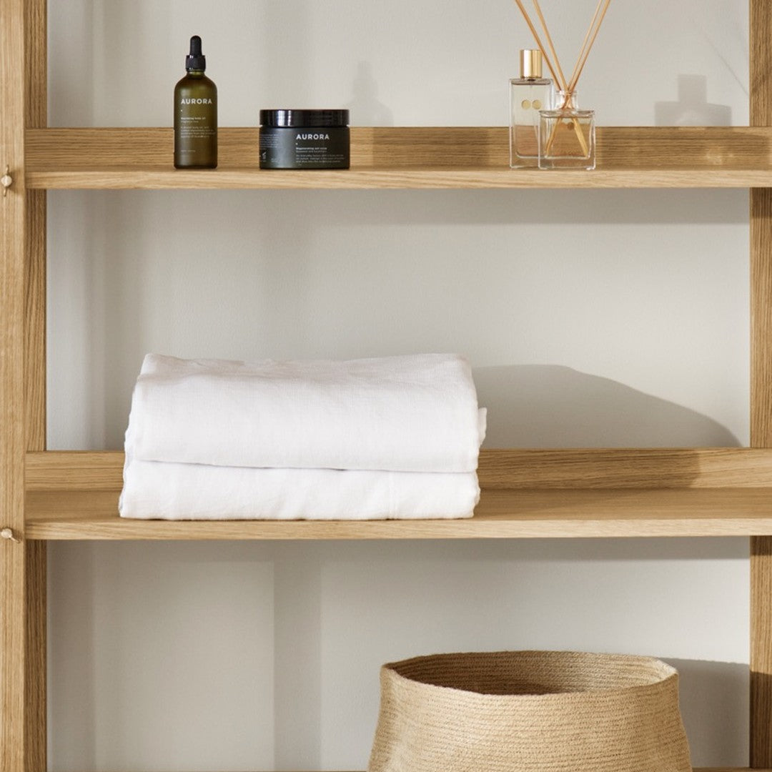 White towels on a light wood shelf, with dark glass bottles and a woven basket in soft natural tones.