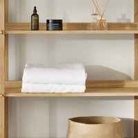 White towels on a light wood shelf, with dark glass bottles and a woven basket in soft natural tones.