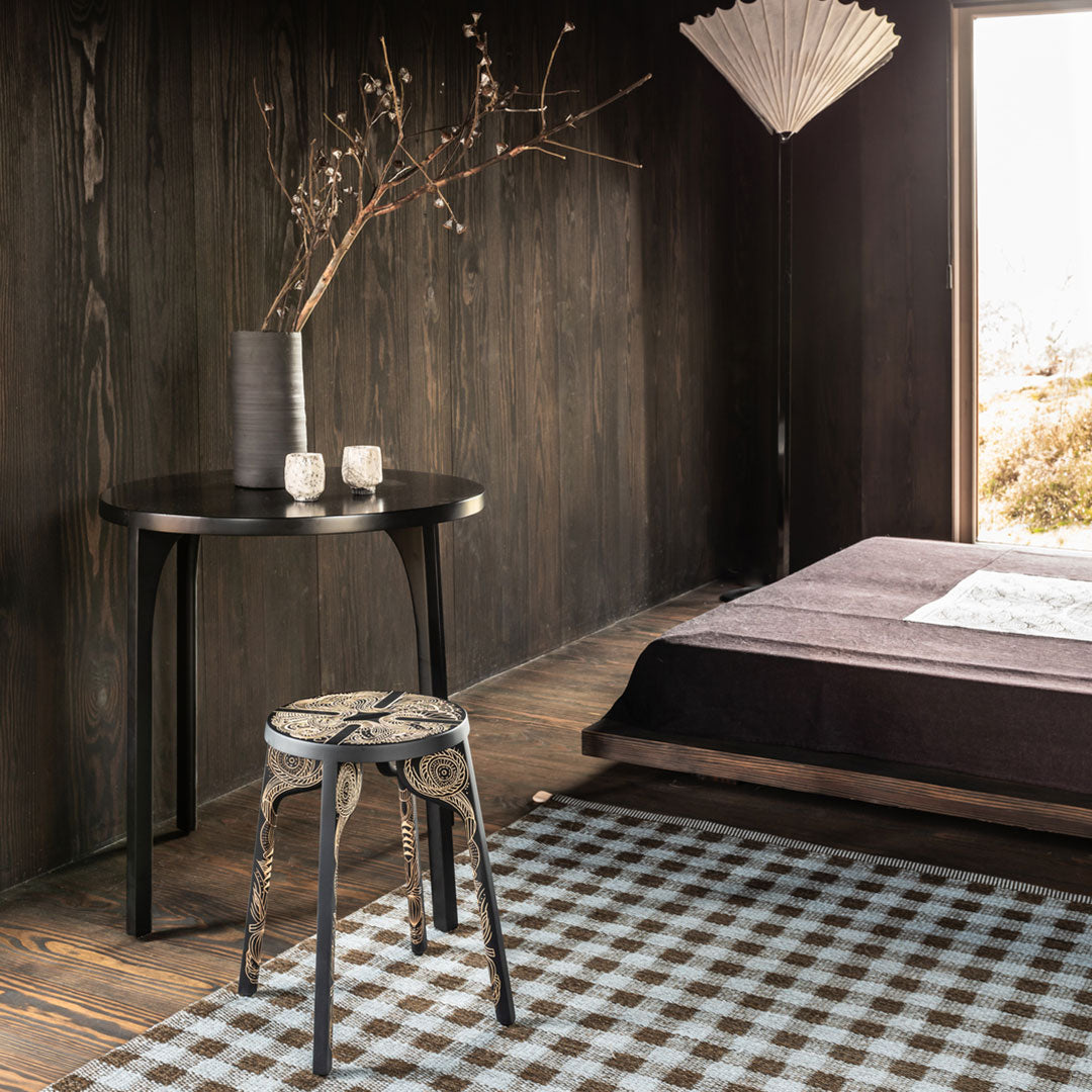 Black wood table and textured gray bedding complement a checkered rug in earthy tones for a cozy, modern aesthetic.