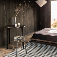 Black wood table and textured gray bedding complement a checkered rug in earthy tones for a cozy, modern aesthetic.