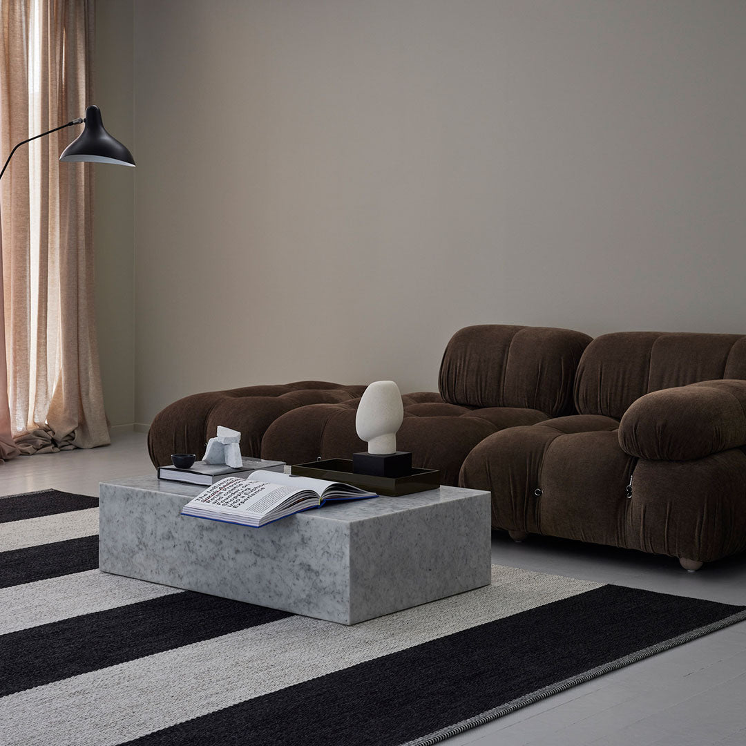 Brown plush sofa, gray stone coffee table, and black and white striped area rug complement a neutral wall backdrop.