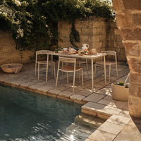 "White outdoor dining set with beige accents, surrounded by natural stone and lush greenery by a blue pool."