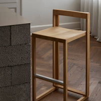 Natural wood bar stool with a minimalist design, featuring a light oak finish and clean lines against a textured backdrop.