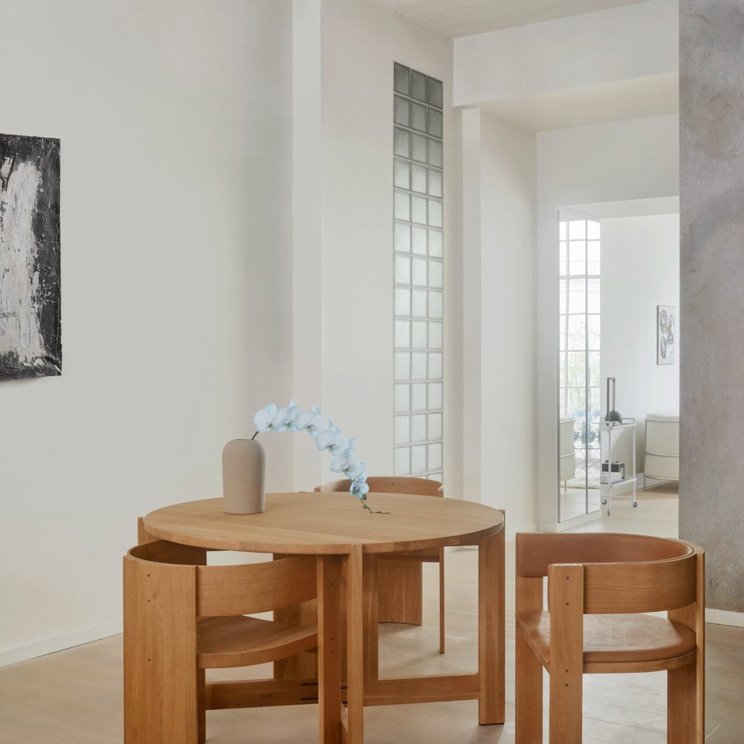Light wood round dining table with natural finish, complemented by minimalist wooden chairs in a soft, neutral-toned interior.