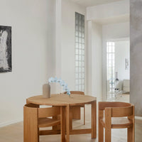 Light wood round dining table with natural finish, complemented by minimalist wooden chairs in a soft, neutral-toned interior.