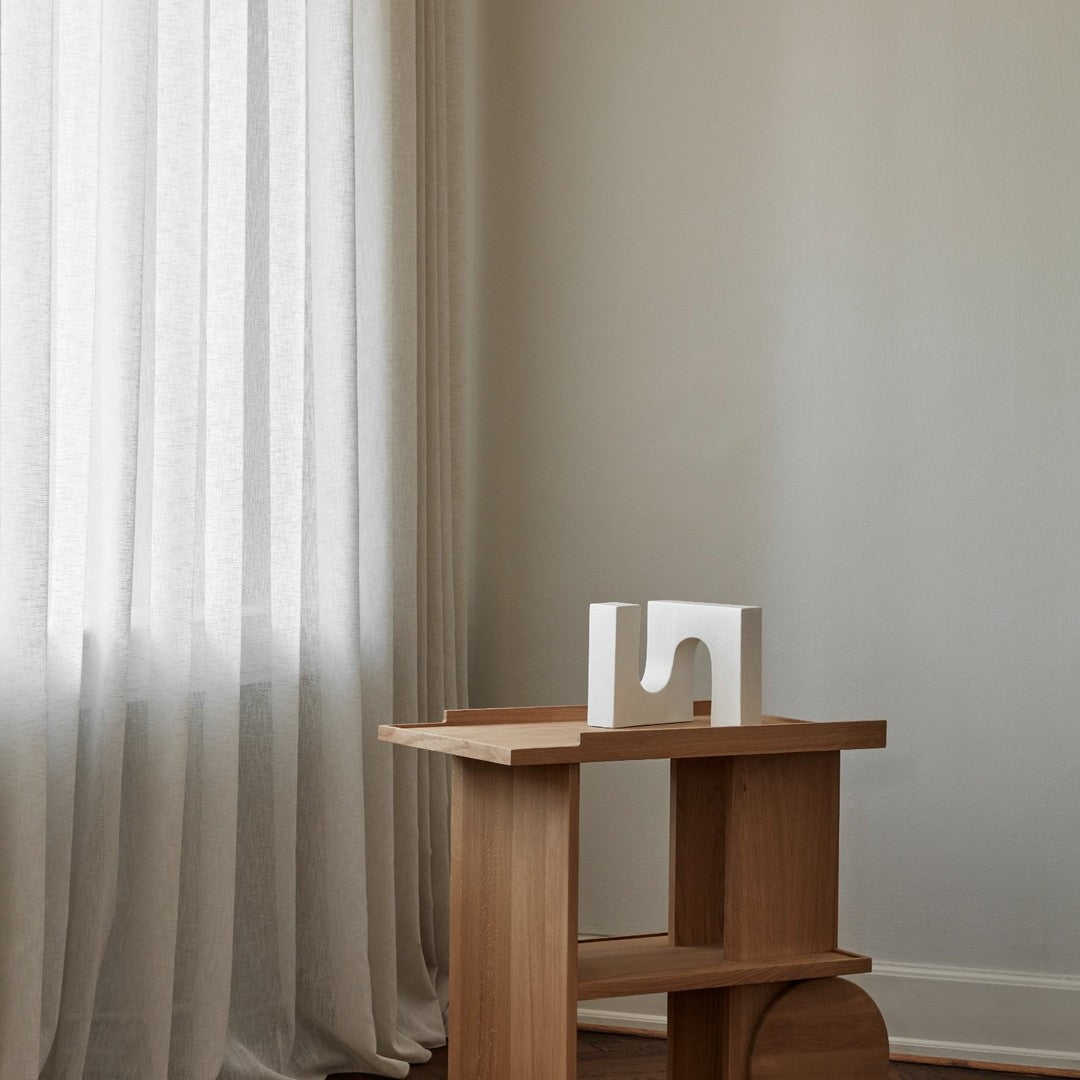 Light wooden side table with a minimalist white decor piece against soft, neutral-toned walls and sheer curtains.