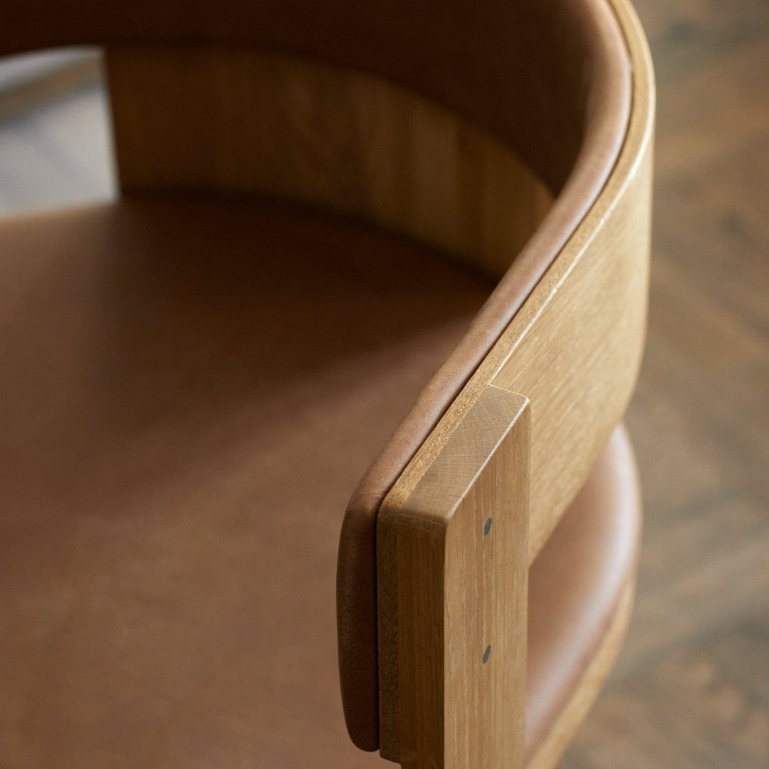 Close-up of a curved wooden chair arm with a warm brown leather seat and smooth natural wood grain pattern.