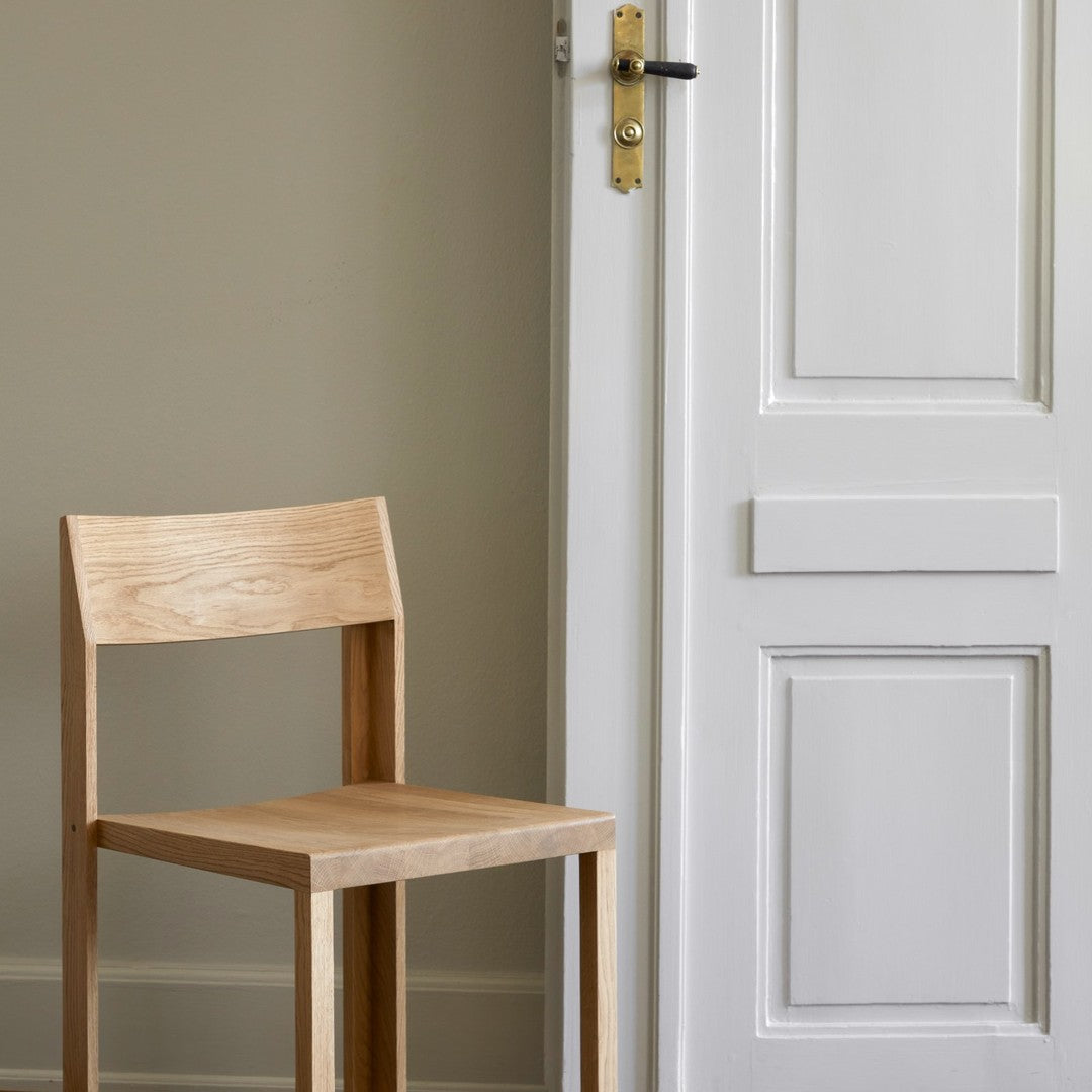 Natural wood chair with a light, warm finish against a neutral textured wall and a white door. Simple and modern design.