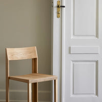 Natural wood chair with a light, warm finish against a neutral textured wall and a white door. Simple and modern design.