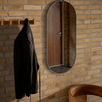 "Modern oval mirror with silver frame, set against warm brick wall and paired with a black jacket and brown chair."