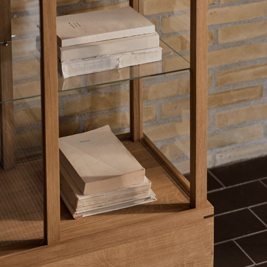 Natural wood cabinet with glass shelves, displaying stacked pale books against a textured stone wall.