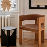 Modern wooden chair with tan leather seat beside a minimalist black side table; warm tones and geometric patterns.