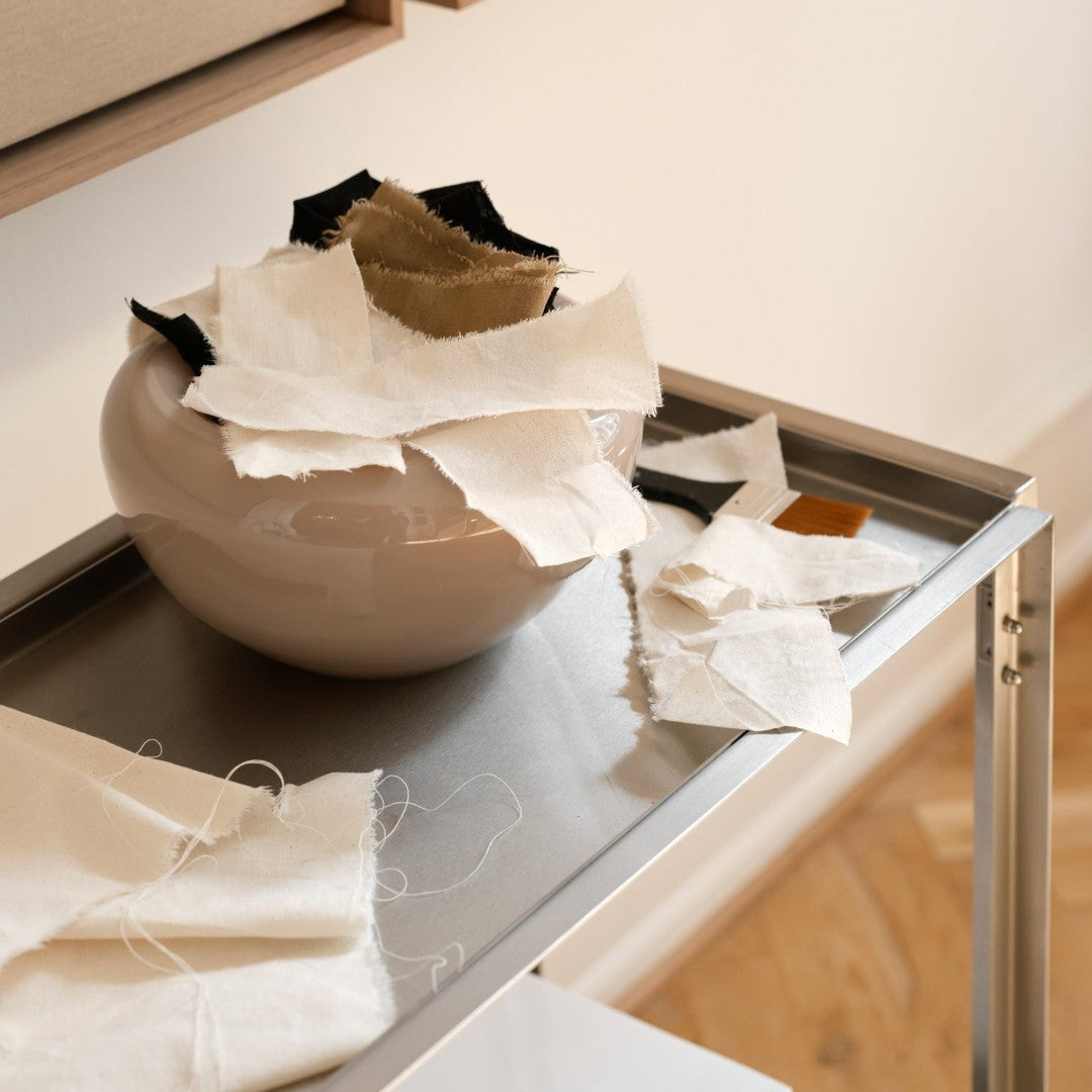 Neutral-toned fabric scraps in a soft, round ceramic bowl, complemented by a sleek metal tray and natural wooden floor.