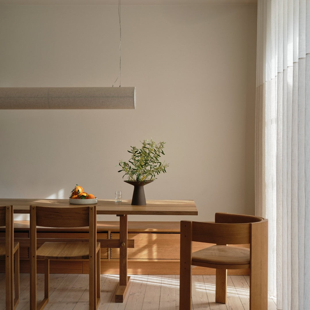 Wooden dining table with natural finishes, simple black vase, and green foliage, complemented by light neutral walls.