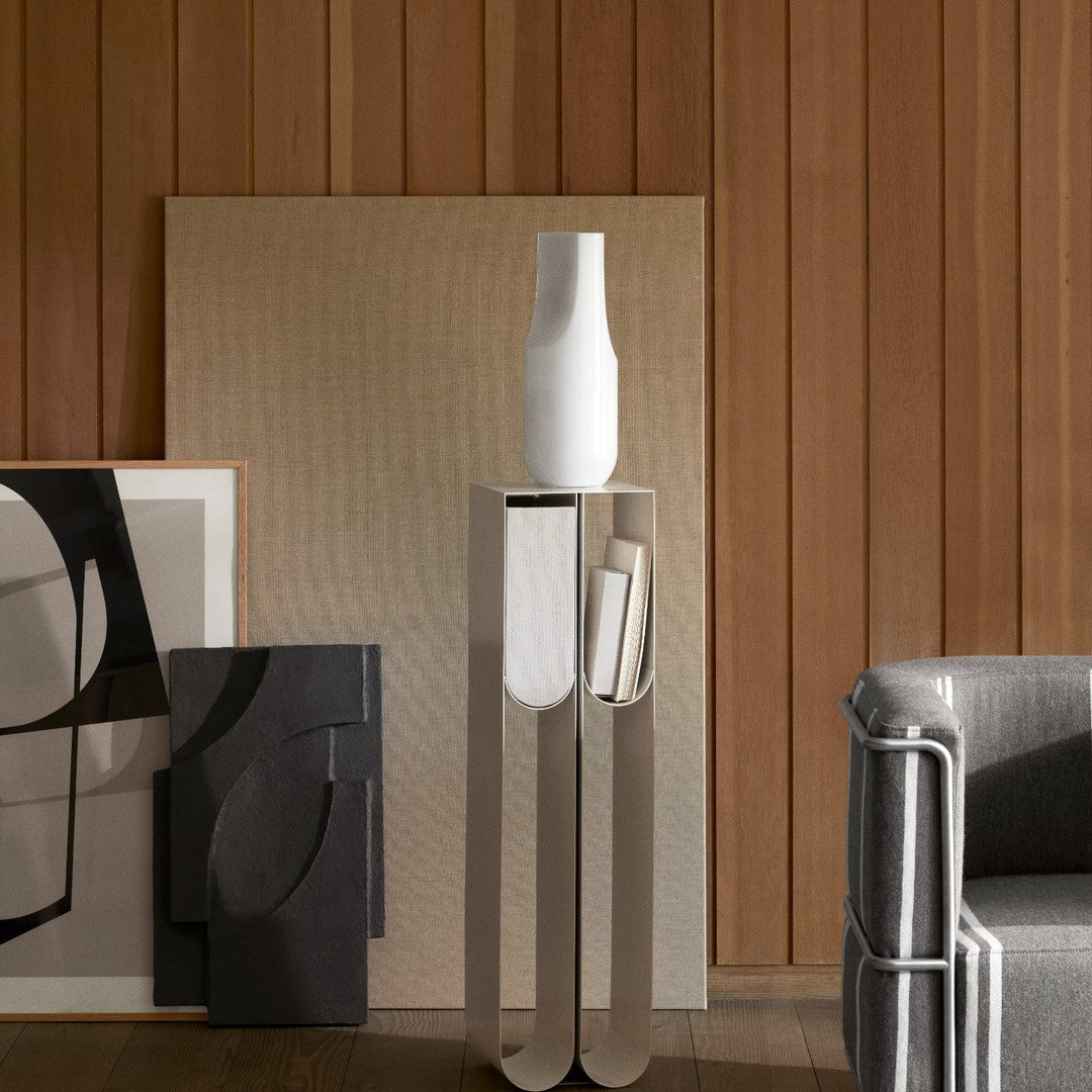 Modern silver pedestal with a white vase, paired with black and white abstract art on warm wooden backdrop.