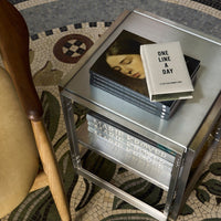 "Silver minimalist side table with stacked books, featuring a patterned rug in earth tones and intricate designs."
