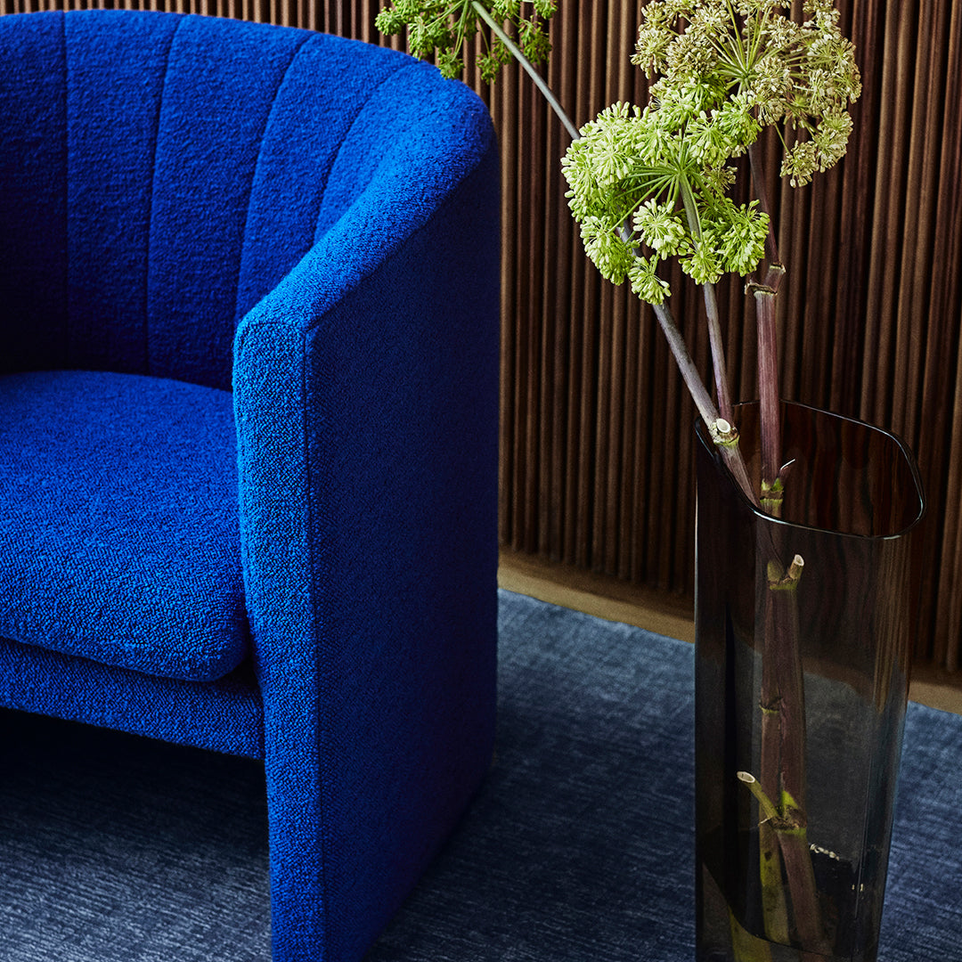 Vibrant blue textured armchair beside a sleek transparent vase, set against a dark blue rug and warm wooden backdrop.