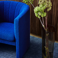Vibrant blue textured armchair beside a sleek transparent vase, set against a dark blue rug and warm wooden backdrop.