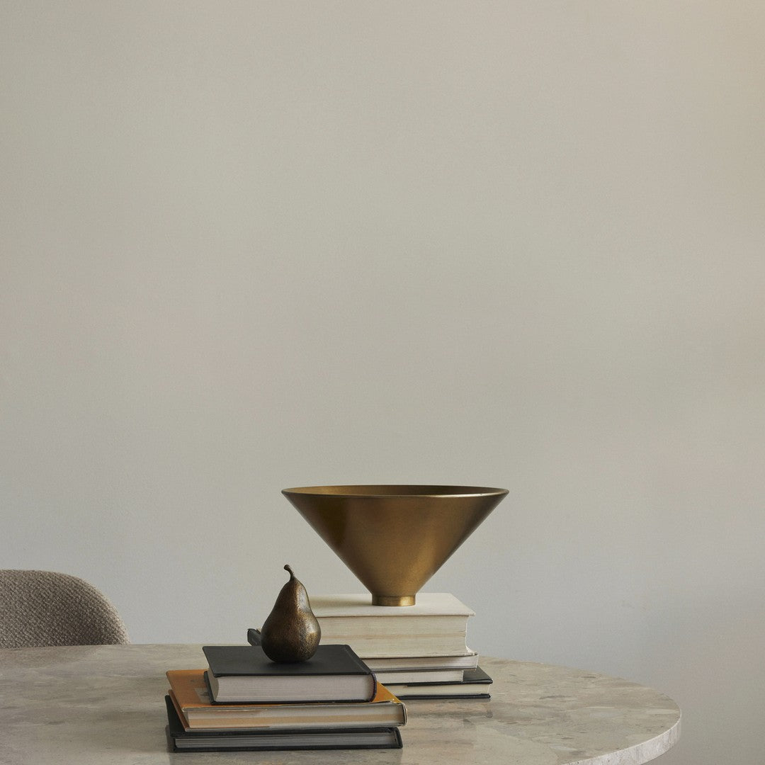 Bronze cone-shaped bowl atop stacked white and black books, with a dark pear accent on a marble table.