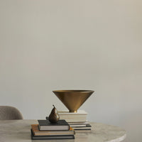 Bronze cone-shaped bowl atop stacked white and black books, with a dark pear accent on a marble table.
