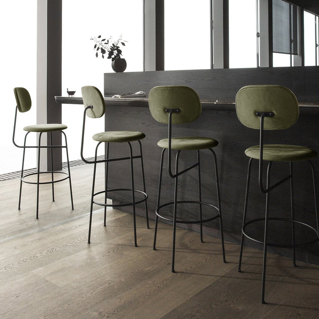 Green upholstered bar stools with black metal frames, set against a minimalistic wooden floor backdrop.