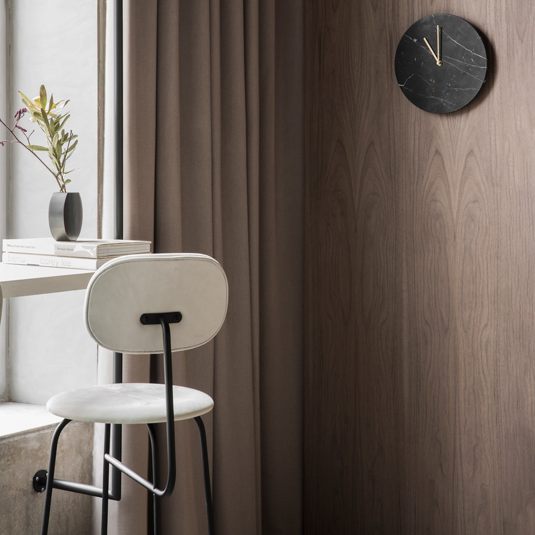 Minimalist design featuring a white chair, dark wood wall, and a black clock, complemented by soft beige curtains.