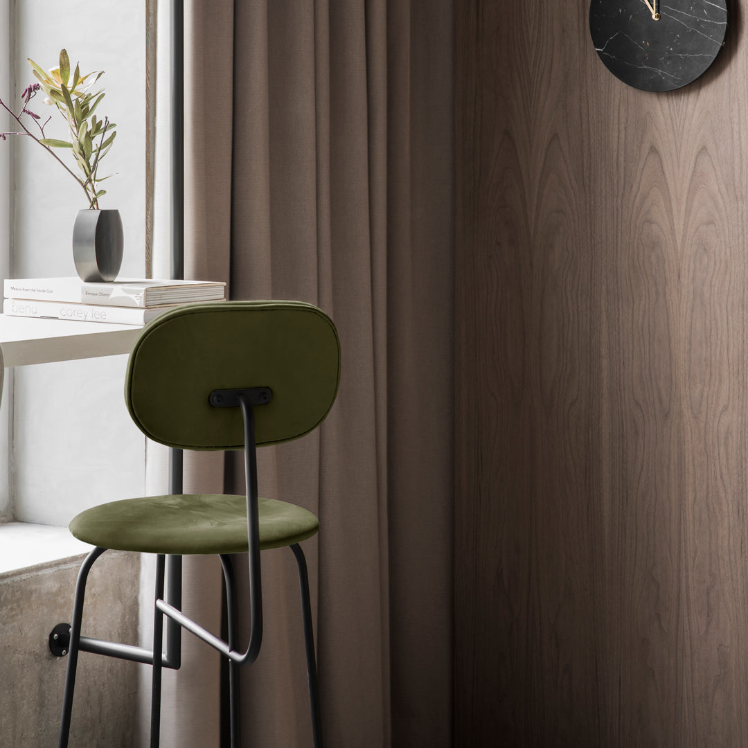 Green upholstered stool with black metal legs, surrounded by warm beige curtain and natural wood grain wall.