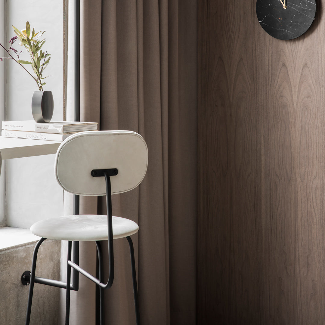 Stylish white bar stool with black metal legs against warm, neutral-toned wooden wall and soft brown curtains.