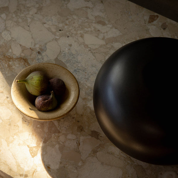 Black ceramic bowl on a textured beige marble surface with a light brown stoneware dish holding green and purple figs.