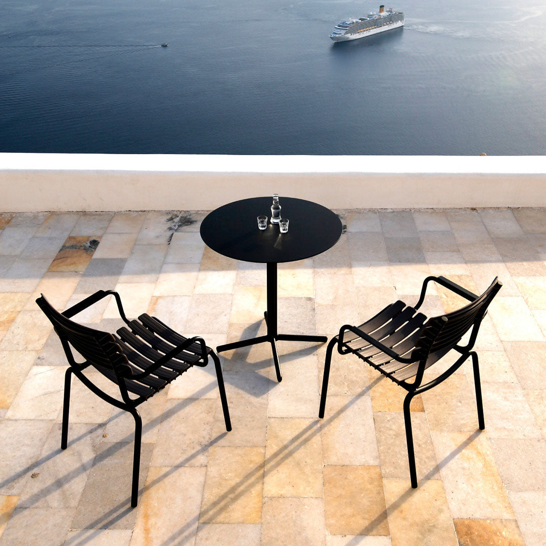 Black striped chairs and a round black table atop a light-toned tiled patio overlooking the water.
