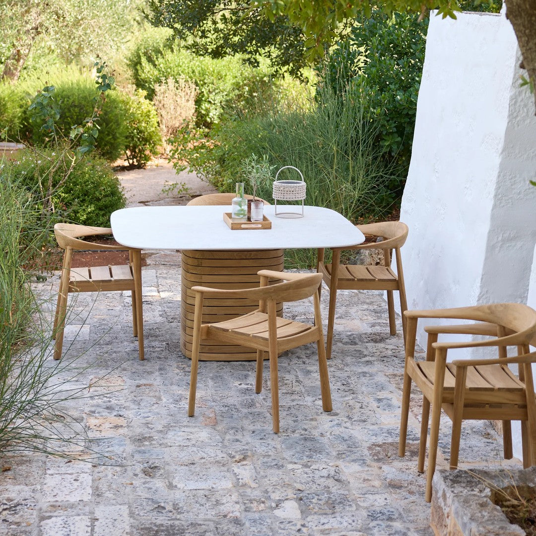 Light wood dining set with a white tabletop, surrounded by lush greenery and stone patio for a serene outdoor vibe.