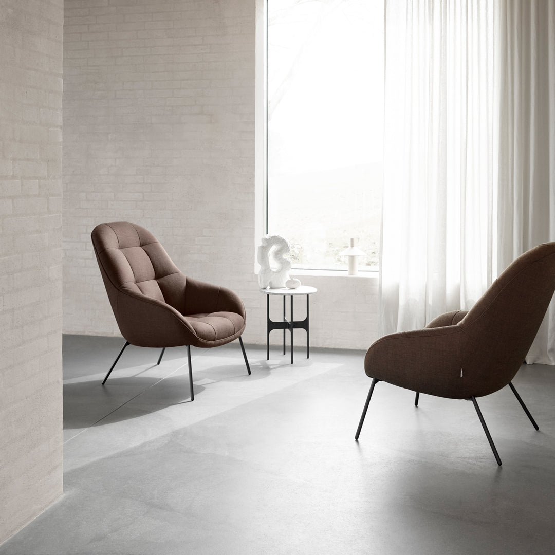 "Plush brown armchairs with sleek black legs, set against a minimalist gray floor and bright, soft curtains."