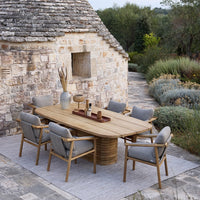 Outdoor dining table with natural wood finish and gray cushions, surrounded by lush greenery and stone textures.