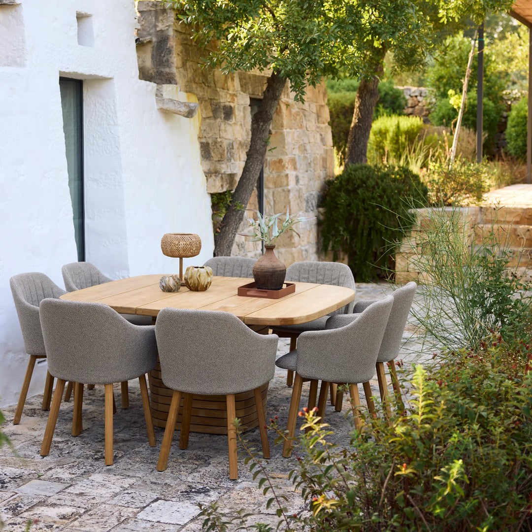 "Natural wood table with light gray upholstered chairs in a serene outdoor setting, surrounded by greenery and stone walls."