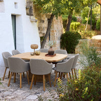 "Natural wood table with light gray upholstered chairs in a serene outdoor setting, surrounded by greenery and stone walls."