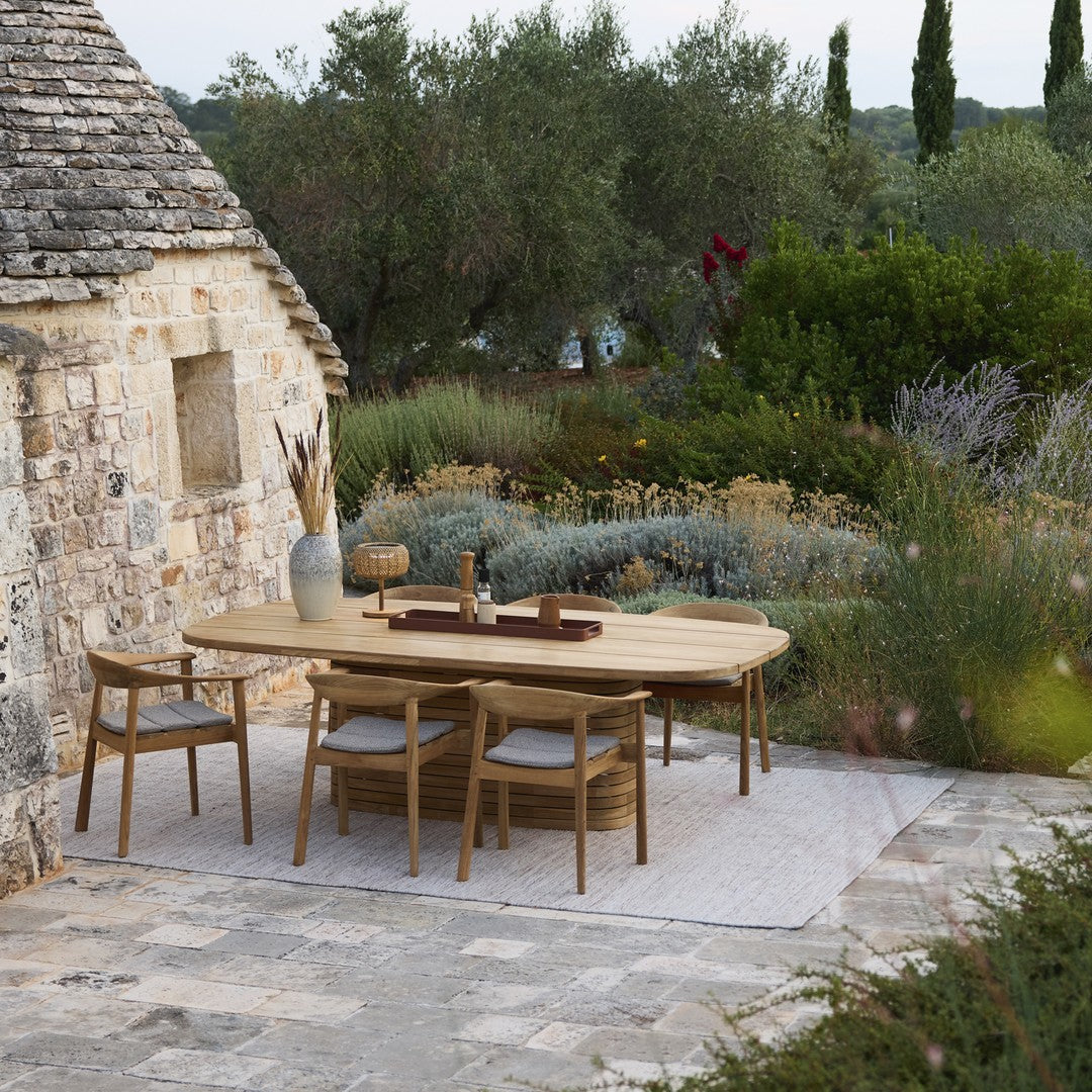 Elegant light wood dining table with natural woven chairs against a backdrop of lush greenery and earthy stone textures.