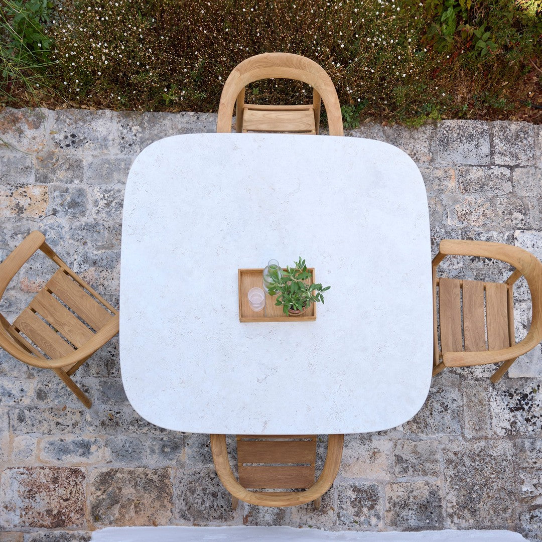 Square white marble table with wooden chairs, adorned by a small green plant centerpiece on a stone patio.