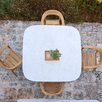 Square white marble table with wooden chairs, adorned by a small green plant centerpiece on a stone patio.