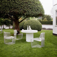Transparent acrylic chairs and a smooth white table set against a lush green lawn and manicured bushes.