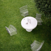 White round table surrounded by transparent mesh chairs on a lush green lawn. Modern design with clean lines and minimalist style.