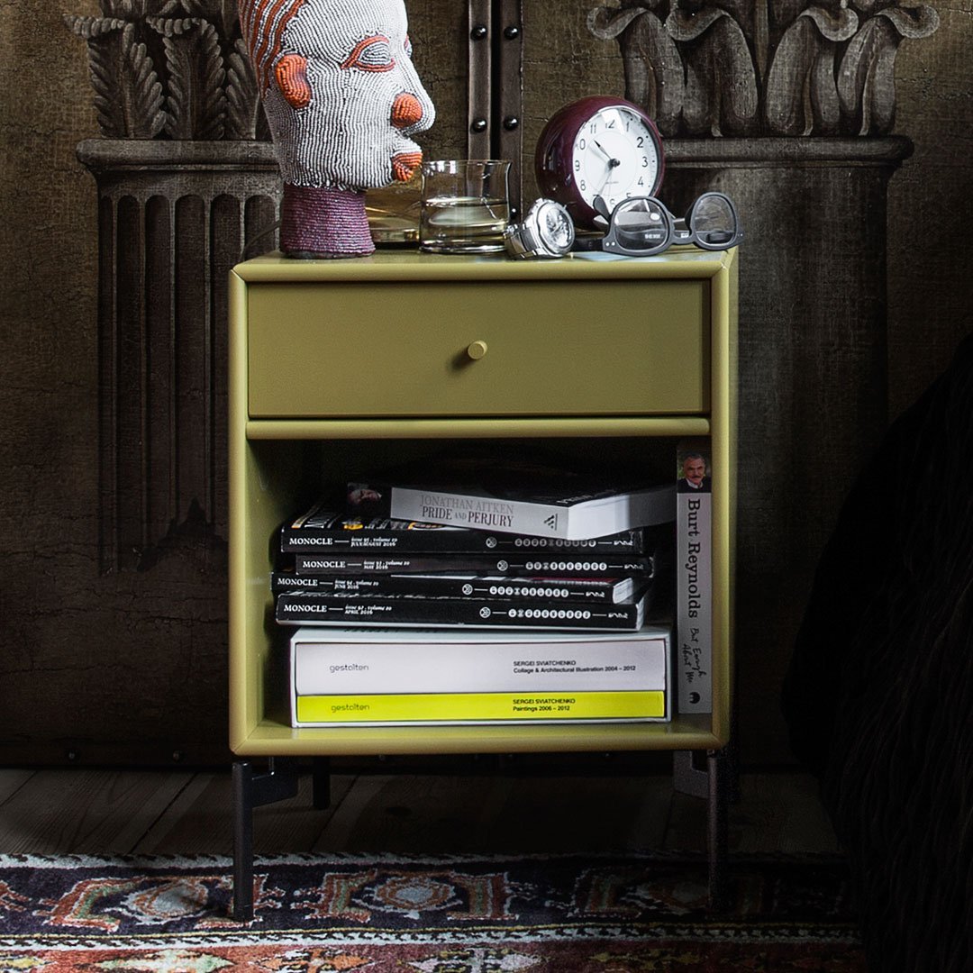 Green nightstand with black legs, orange and gray decor accent, stacked monochrome magazines, and a patterned rug.
