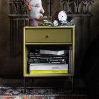 Green nightstand with black legs, orange and gray decor accent, stacked monochrome magazines, and a patterned rug.