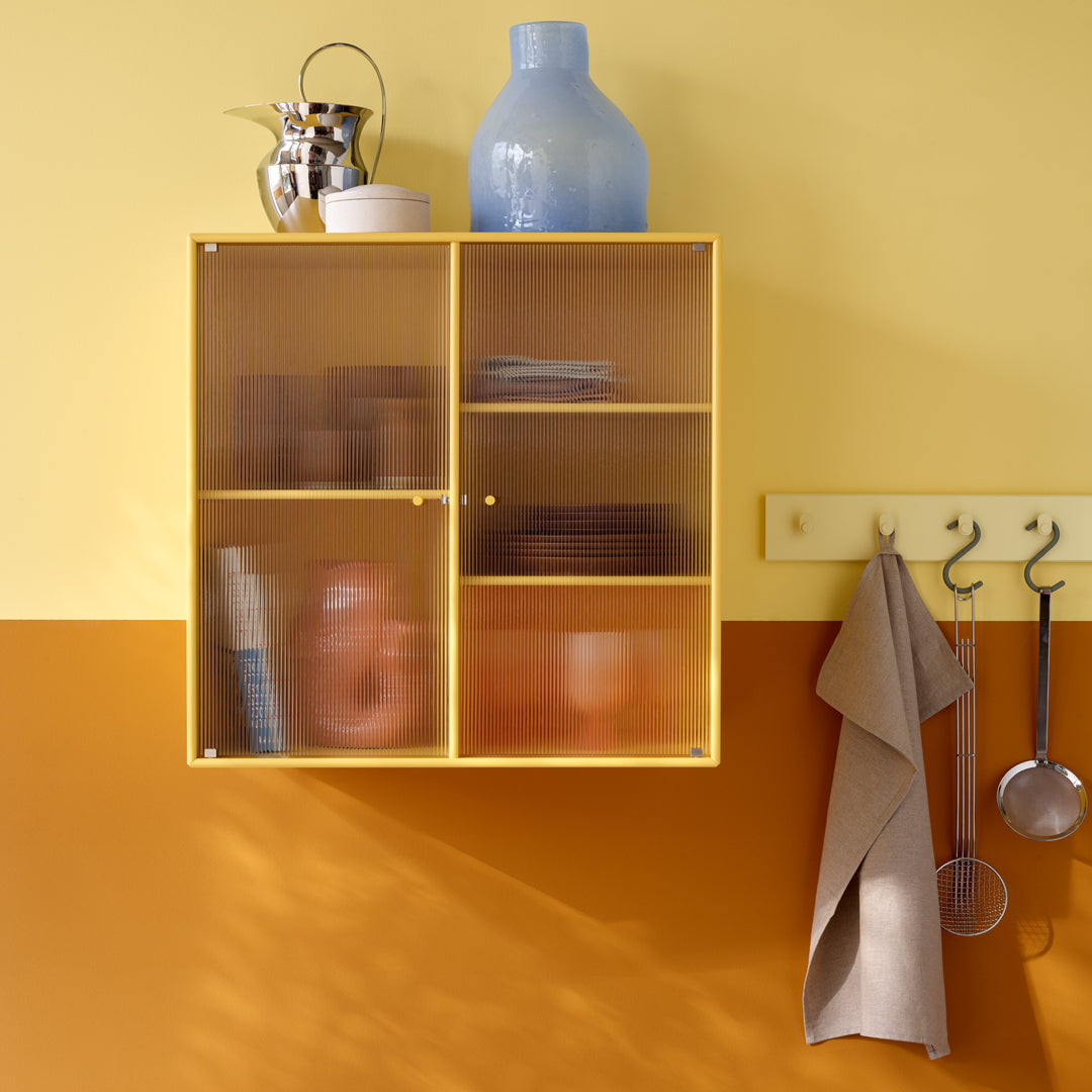 Yellow mesh cabinet against a soft yellow wall, with a warm orange lower section and blue vase on top.