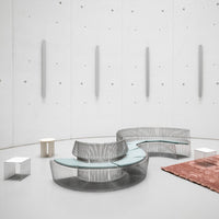 Modern curved coffee table in silver metal with a glass top, set on a gray floor with a textured orange rug.