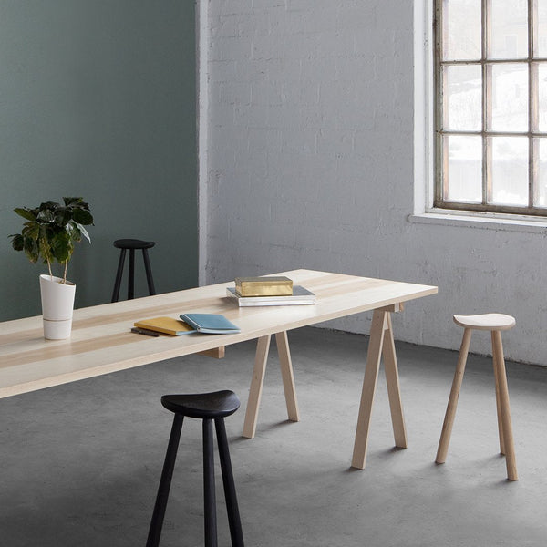 Light wood table with natural finish, complemented by black and white stools on a gray floor and green wall.