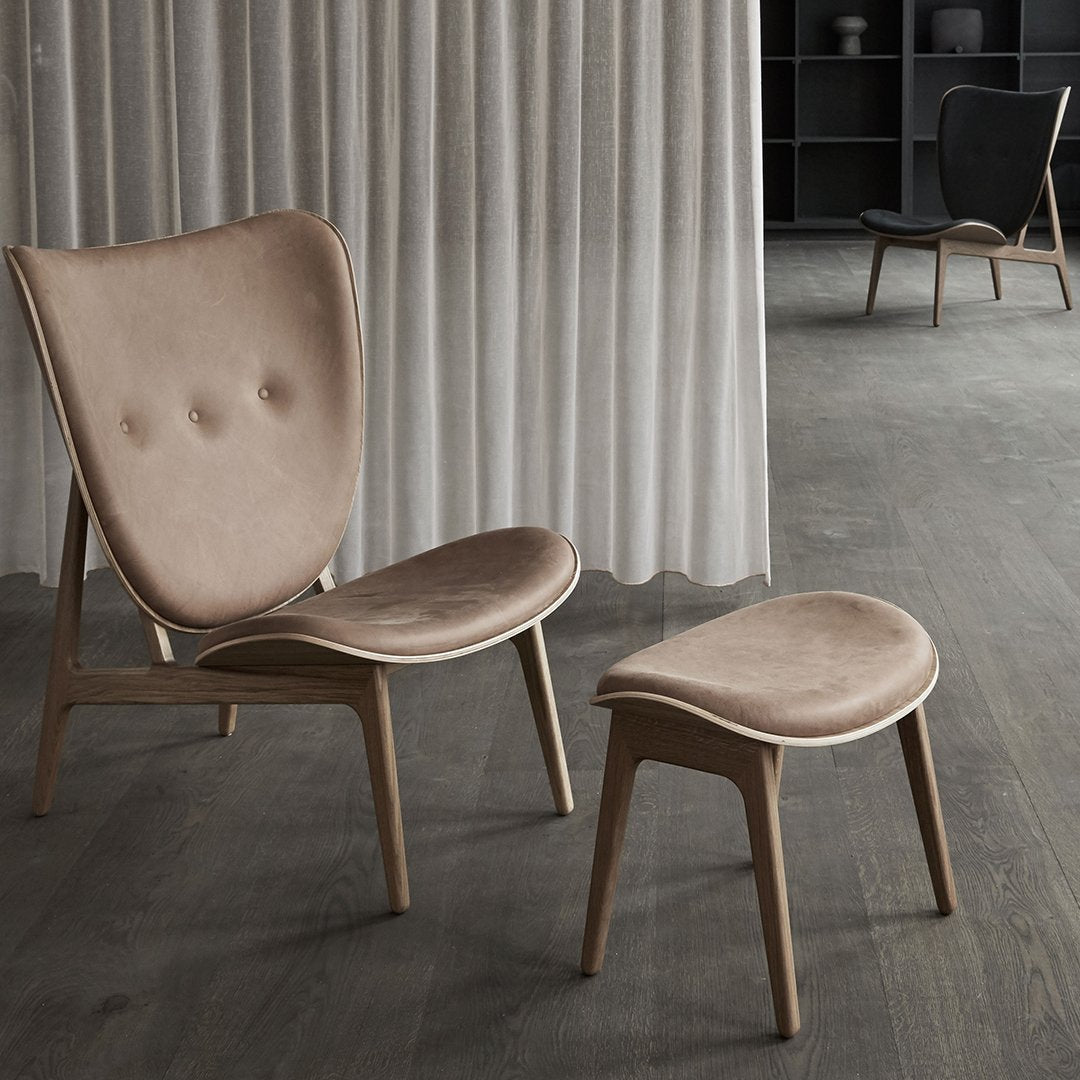 "Beige velvet lounge chair and matching ottoman on dark wooden floor with soft curtains in background."
