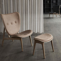 "Beige velvet lounge chair and matching ottoman on dark wooden floor with soft curtains in background."