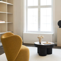 Mustard yellow armchair with smooth curves, contrasted by a round black table, set against a neutral beige backdrop.